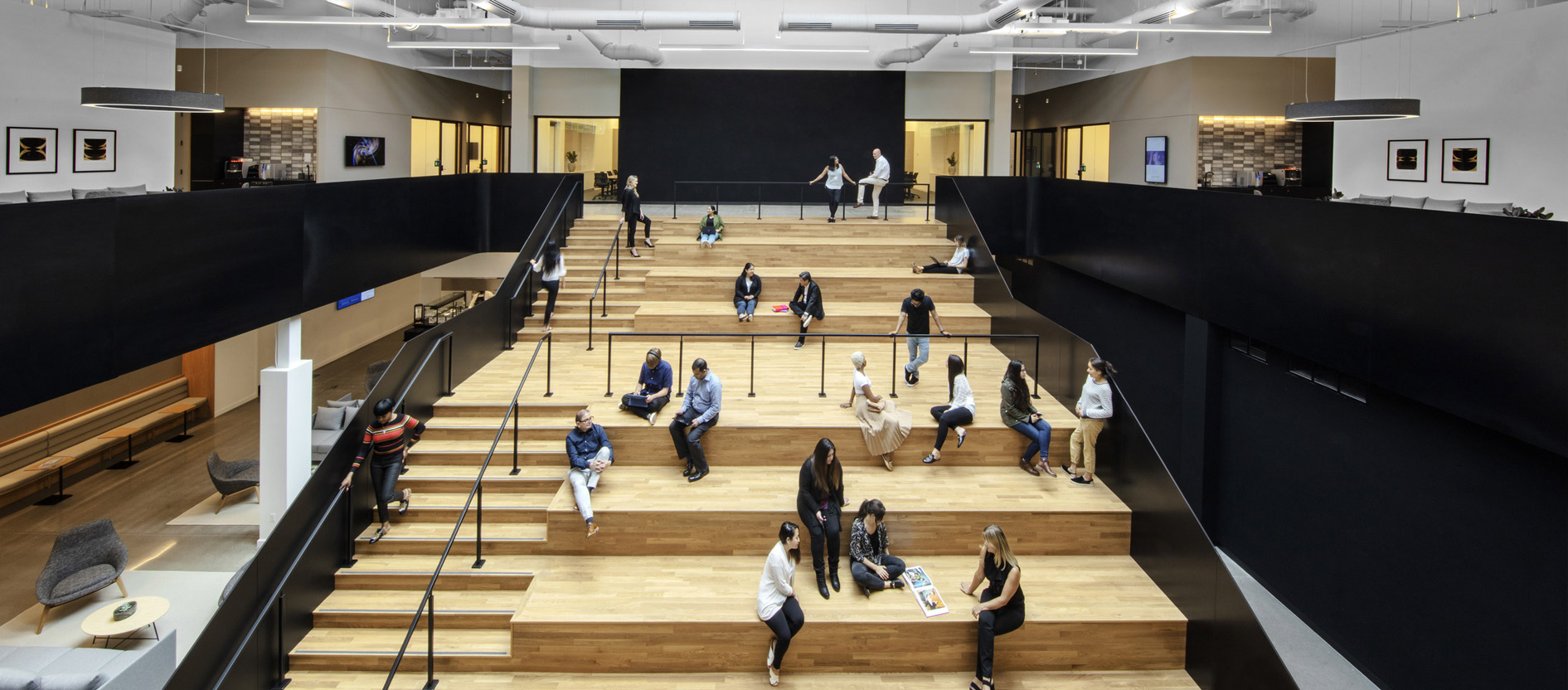 Modern office common area with employees collaborating and relaxing on tiered wooden seating steps, surrounded by sleek architectural features and ample natural light.