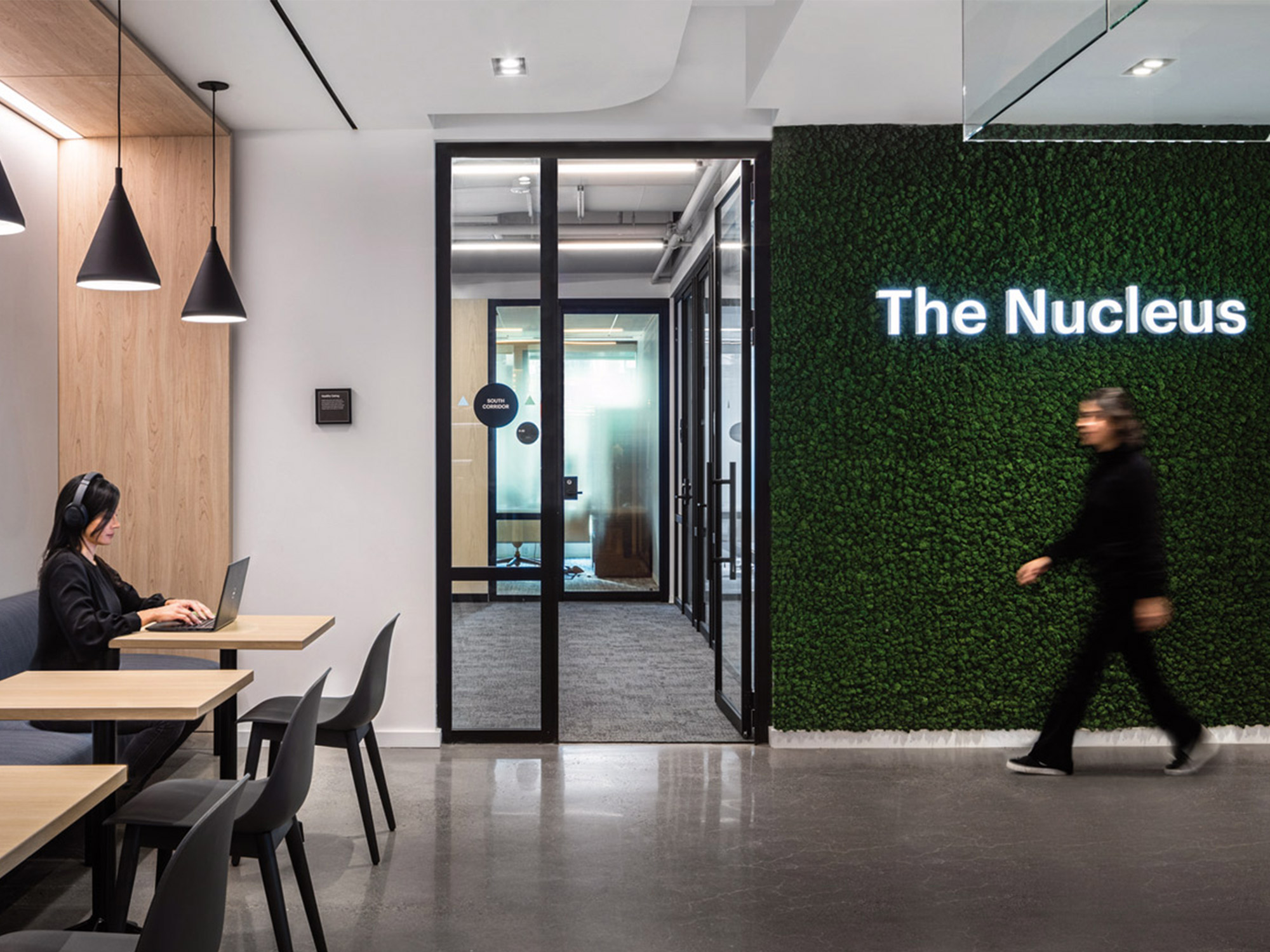 A modern office environment featuring a sleek workspace with an individual working at a long table, pendant lights above, and a vibrant green living wall with the label "the nucleus" illuminated, delineating a space for focused work or collaboration.