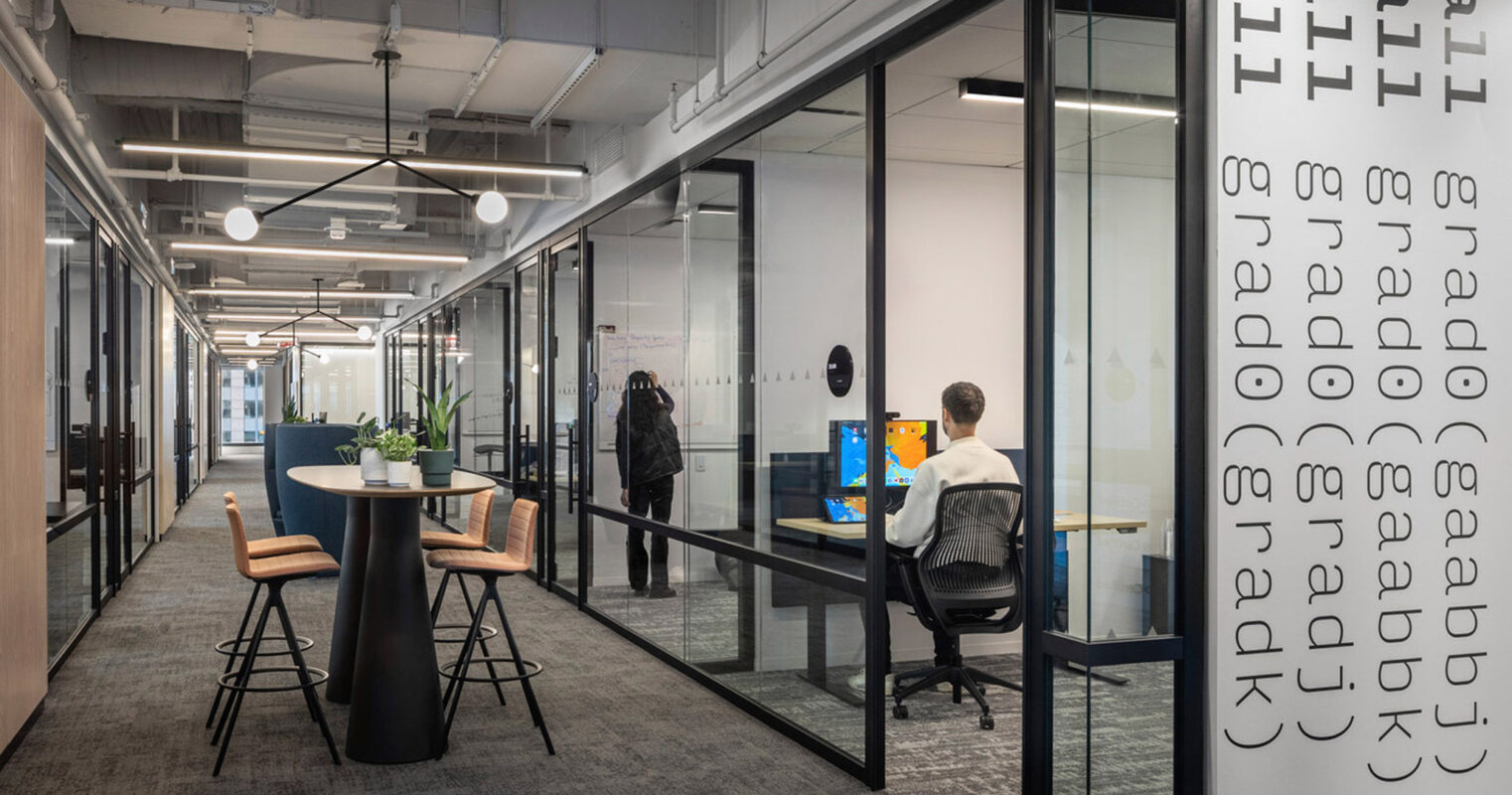 Modern office interior featuring exposed ceiling pipes, linear pendant lighting, and glass-walled meeting rooms. Bar-height tables pair with stools for casual workspaces. Subtle branding appears on the glass with frosted lettering.